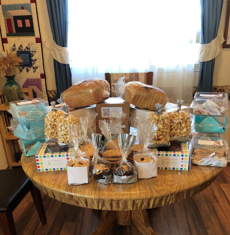 May include: A table filled with baked goods, including loaves of bread, popcorn, cookies, and brownies. The table is covered in a brown tablecloth and has a variety of containers and bags holding the food.