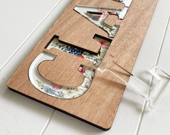 May include: A wooden sign with the word "Clara" cut out, revealing a floral pattern underneath. The sign is mounted on a clear acrylic stand.