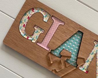 May include: A wooden sign with the word "GLAM" cut out and decorated with patterned paper. The letters are in different colors and patterns, including pink, blue, and white.