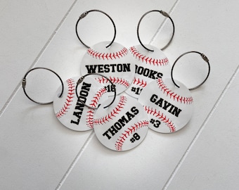 May include: Five white baseball-themed keyrings with black lettering and red stitching. The keyrings feature the names "Landon", "Weston", "Brooks", "Thomas", and "Gavin" with corresponding numbers.