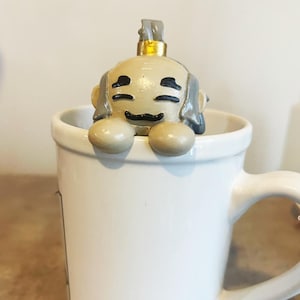 May include: A white ceramic mug with a small, gray and beige figurine of a character with a gold crown sitting on the rim.