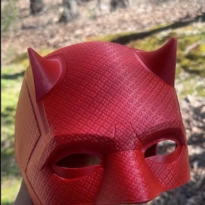 May include: A red plastic Daredevil mask with a textured surface and two horns. The mask has eye holes and a raised design on the forehead.