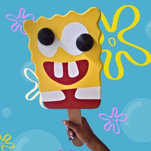Może przedstawiać: Żółta, prostokątna dekoracja ścienna w kształcie loda na patyku SpongeBob Kanciastoporty z drewnianym uchwytem. Posiada duże białe oczy z czarnymi źrenicami, szerokie czerwone usta z dwoma białymi zębami oraz białą koszulę nad czerwonymi spodniami.