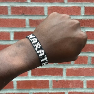 May include: A black and white beaded bracelet with a geometric pattern. The bracelet is worn on a person's wrist.