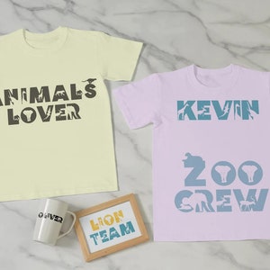 Peut inclure: Deux t-shirts de couleur claire avec des silhouettes d'animaux qui &eacute;pellent "Animals Lover" et "Kevin Zoo Crew". Une tasse et une pancarte en bois avec le texte "Lion Team" sont &eacute;galement pr&eacute;sents sur l'image.