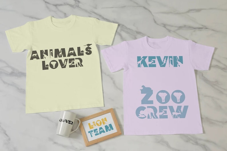 Peut inclure: Deux t-shirts de couleur claire avec des silhouettes d'animaux qui &eacute;pellent "Animals Lover" et "Kevin Zoo Crew". Une tasse et une pancarte en bois avec le texte "Lion Team" sont &eacute;galement pr&eacute;sents sur l'image.