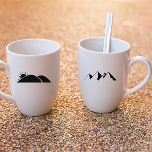 Puede incluir: Dos tazas de cer&aacute;mica blanca con dise&ntilde;os gr&aacute;ficos negros. Una taza tiene un dise&ntilde;o de cordillera y la otra tiene un dise&ntilde;o de sol saliendo sobre una colina.