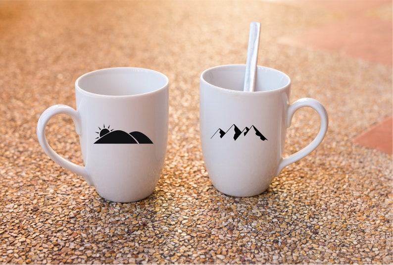 Puede incluir: Dos tazas de cer&aacute;mica blanca con dise&ntilde;os gr&aacute;ficos negros. Una taza tiene un dise&ntilde;o de cordillera y la otra tiene un dise&ntilde;o de sol saliendo sobre una colina.