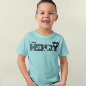Peut inclure: Un t-shirt bleu clair avec un motif noir du nom "HENRY" &eacute;crit en silhouettes d'animaux. Les silhouettes comprennent un panth&egrave;re, un oiseau, un &eacute;l&eacute;phant et un ours.