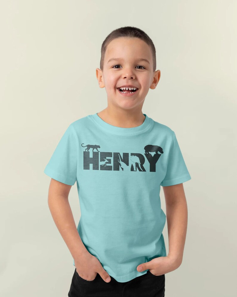 Peut inclure: Un t-shirt bleu clair avec un motif noir du nom "HENRY" &eacute;crit en silhouettes d'animaux. Les silhouettes comprennent un panth&egrave;re, un oiseau, un &eacute;l&eacute;phant et un ours.