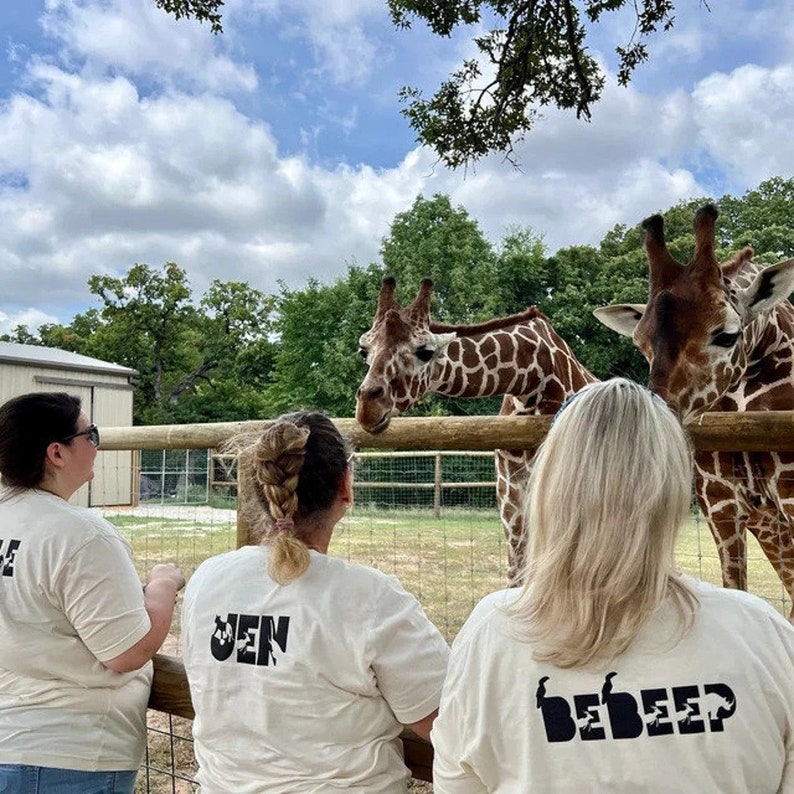 Peut inclure: Trois personnes portant des t-shirts blancs avec du texte noir se tiennent devant une cl&ocirc;ture regardant deux girafes. Le texte sur les t-shirts est "Jen", "Be" et "Deep".