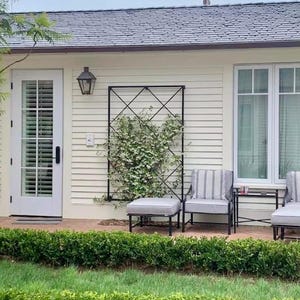 May include: An outdoor patio setting featuring a black metal trellis with climbing plants. Two chairs with striped cushions and an ottoman are on a brick patio. A white door and window are visible, along with a black lantern.