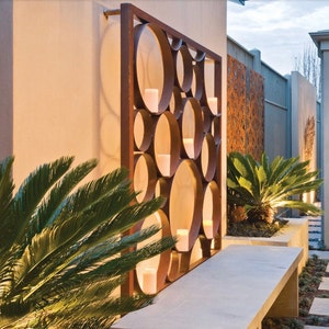 May include: A contemporary outdoor area with a metal wall sculpture featuring a geometric pattern of circles. The sculpture is lit by warm light, creating a cosy and inviting atmosphere. A stone bench sits in front of the sculpture, providing a place to relax and enjoy the surroundings.