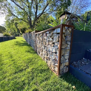 Könnte beinhalten: Eine Gabionenmauer, gefüllt mit Steinen und umrahmt von einer rostigen Metallstruktur, steht in einem grasbewachsenen Garten. Die Mauer ist mit unterschiedlich großen Steinen gefüllt. Der Hintergrund umfasst grünes Gras, Bäume und einen blauen Himmel.