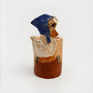 Könnte beinhalten: Keramik-Entenfigur mit blauer Haube, die aus einem zylindrischen Topf herausragt. Die Ente hat einen beigen Körper, einen orangefarbenen Schnabel und ist vor einem weißen Hintergrund platziert. Der Topf ist beige mit braunem Boden und grünen Akzenten.