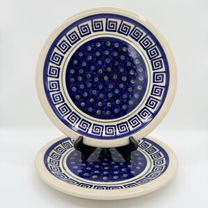 May include: Two ceramic plates with a blue and white design. The plates have a cream-colored rim and a blue center with a pattern of small green dots. A border of blue geometric shapes surrounds the center. The plates are approximately 9 inches in diameter.