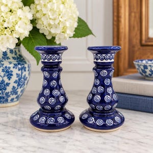 May include: Two matching ceramic candle holders featuring a deep blue glaze with a repeating white spiral pattern. Each holder has a wide, rounded base, a curved neck, and a flared top rim, designed to securely hold a single taper candle.
