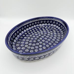 May include: Oval ceramic baking dish with a dark blue base and white spiral pattern. The rim features a decorative border of white swirls and dots. This dish is ideal for serving or baking.