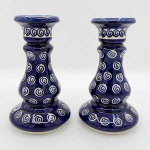 May include: Two matching ceramic candle holders. The candle holders are navy blue with white spiral and swirl patterns. The base and top of the candle holders are a lighter shade of blue. The candle holders are approximately 6 inches tall.