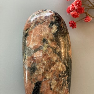 May include: A smooth, polished, oval-shaped stone with a unique pattern of black, pink, and white. The stone has a natural, organic look.