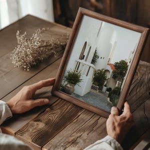 Peut inclure: Un cadre photo en bois avec un passe-partout blanc et une photographie d'un bâtiment blanc avec une cour. Le bâtiment a un toit en tuiles et un balcon avec une balustrade. Il y a des plantes dans la cour.