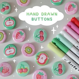 May include: A collection of hand-drawn buttons with fruit designs, including strawberries, cherries, apples, and kiwi. The buttons are in shades of pink and green. The text "HAND DRAWN BUTTONS" is in a white cloud-shaped text box. Colored markers are in the background.