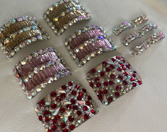 May include: A collection of rectangular and curved metal hair clips adorned with various colored rhinestones. The set includes pieces with gold, pink, and red crystals, all bordered by clear, iridescent stones that catch the light.