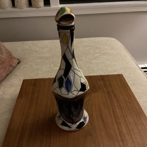 May include: A tall, decorative ceramic bottle with a geometric pattern. The bottle features a white background with black, blue, yellow, and green diamond shapes. The stopper is multi-colored. The bottle is displayed on a wooden surface.