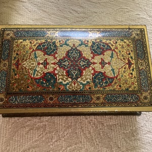 May include: Rectangular gold-toned metal box with an ornate, colorful design. The lid features a central floral pattern in teal, red, and cream, surrounded by Arabic script. The box has a vintage aesthetic.