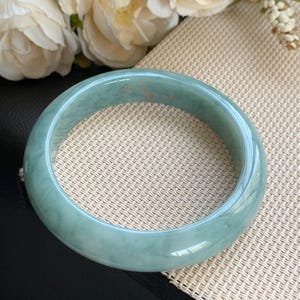 May include: A smooth, polished jade bangle bracelet featuring a translucent light green color with subtle darker green veining and small, faint golden-brown inclusions throughout the circular band, showcasing a glossy, reflective surface finish.
