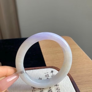 May include: A smooth, circular bangle bracelet crafted from translucent stone, featuring a soft lavender-purple hue that transitions into a milky white tone. The polished surface of the bangle reflects light, highlighting its rounded, solid form.