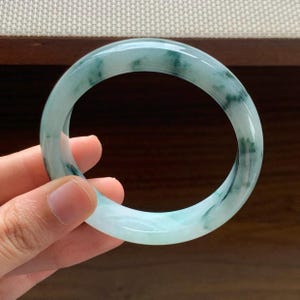 May include: A smooth, circular bangle bracelet crafted from translucent pale green stone. The surface features distinct, irregular dark green mineral inclusions and mottled patterns throughout the polished material, creating a natural, variegated appearance.