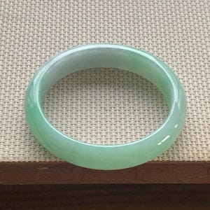 May include: A smooth, circular bangle bracelet crafted from translucent light green jade stone. The piece features a polished, rounded surface with subtle variations in colour intensity, ranging from pale mint to soft emerald hues throughout the band.
