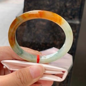 May include: A smooth, circular bangle bracelet crafted from polished stone, featuring a gradient of vibrant orange and deep red hues transitioning into a translucent, pale mint green. A small, thin red string is tied around the lower section.