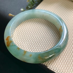 Op de afbeelding: Een gladde, gepolijste jade armband met een doorschijnende lichtgroene basiskleur en opvallende, onregelmatige vlekken van goudbruine en donkergroene mineraalinsluitingen verspreid over de gehele cirkelvormige, massieve stenen bandstructuur.