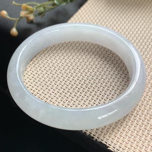 May include: A smooth, polished, translucent white jade bangle bracelet with subtle cloudy inclusions and a rounded profile. The circular jewellery piece features a glossy finish that reflects light, showcasing the natural stone texture and pale colour.