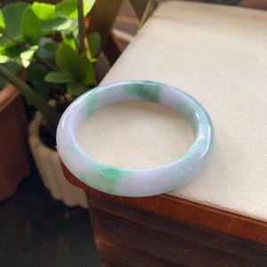 May include: A smooth, circular bangle bracelet crafted from polished stone, featuring a translucent white base with distinct patches of vibrant green and soft lavender hues throughout the rounded surface of the jewellery piece.