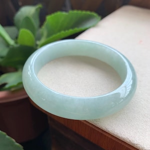 May include: A smooth, polished bangle bracelet crafted from translucent pale green jade. The circular jewellery piece features a rounded profile with a glossy, reflective surface that catches the light, showcasing a consistent light mint green hue throughout.