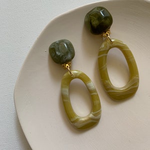May include: A pair of olive green and white marbled oval drop earrings with gold-coloured hardware. The earrings have a square stud top and a large oval shape. The earrings are displayed on a white, slightly textured surface.