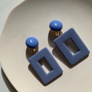 Könnte beinhalten: Ein Paar blaue Statement-Ohrringe in quadratischer Form mit einem Ausschnitt in der Mitte. Die Ohrringe sind aus glattem, mattem Material gefertigt und haben einen goldfarbenen Clipverschluss.