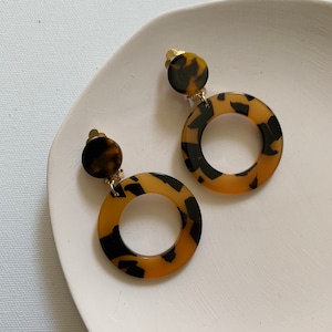 May include: A pair of gold tone clip-on earrings with a brown and black tortoise shell pattern. The earrings feature a large round hoop with a smaller round disc attached to the top.