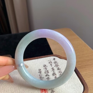 May include: A smooth, circular jade bangle featuring a gradient of translucent pale green and soft lavender hues. The polished stone surface displays natural variations in color and texture throughout the band, creating a continuous, rounded form.