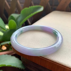 May include: A smooth, circular bangle bracelet crafted from polished stone. The piece features a translucent appearance with a marbled colour palette consisting of soft lavender, pale green, and white tones blended throughout the solid, rounded band.