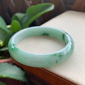 May include: A smooth, circular bangle bracelet crafted from translucent pale green stone. The material features natural, irregular darker green inclusions and veining throughout the polished surface, creating a marbled appearance with a glossy finish.