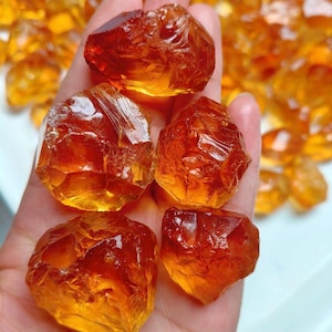 Puede incluir: Una colección de piedras en bruto, translúcidas y de color ámbar con superficies irregulares y facetadas. Estas piedras presentan un tono naranja dorado intenso y una textura similar al vidrio, reflejando la luz en sus bordes dentados y superficies desiguales.
