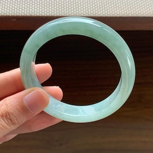 May include: A smooth, polished bangle bracelet crafted from translucent light green jade. The circular jewelry piece features a rounded profile with subtle variations in color intensity and internal cloud-like patterns throughout the stone material.