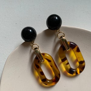 May include: A pair of dangle earrings featuring black round beads and oval-shaped tortoise shell-patterned resin pieces with gold-toned metal accents.