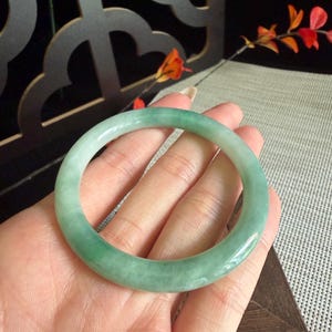 May include: A smooth, circular bangle bracelet crafted from translucent light green jadeite. The piece features a polished, rounded surface with subtle variations in colour intensity and natural internal cloud-like inclusions throughout the stone.