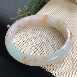 Puede incluir: Un brazalete rígido de piedra blanca translúcida y lisa, con inclusiones irregulares de color naranja rojizo dispersas por toda la superficie pulida. La pieza de joyería circular tiene un perfil redondeado y un acabado brillante con variaciones de color.