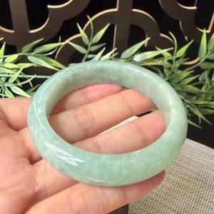 May include: A smooth, polished jade bangle bracelet featuring a translucent, pale green color with subtle white and darker green mottled inclusions throughout the circular stone band, showcasing a glossy finish and a rounded, thick profile.